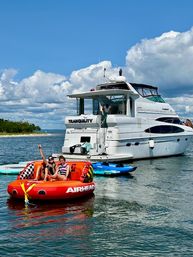 Two people on a bright red Airhead inflatable towable near a white yacht anchored in calm coastal waters with paddleboards nearby under a blue sky with puffy clouds — summer boating scene.