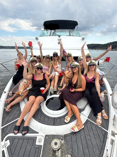 Group of women wearing captain hats and sunglasses on the bow of a white yacht on a lake, raising red cups in a summer yacht party