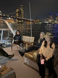Nighttime river cruise: three people relaxing on a boat deck with a lit suspension bridge and glowing city skyline reflecting on the water.