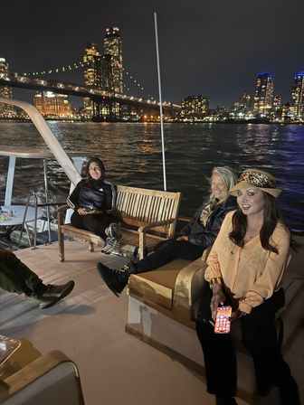Nighttime river cruise: three people relaxing on a boat deck with a lit suspension bridge and glowing city skyline reflecting on the water.
