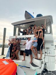 Smiling group of adults on a white yacht docked at a marina, posing and waving with a man in a captain’s hat and an orange inflatable tube visible — lively summer boat party scene.