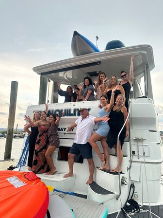 Smiling group of adults on a white yacht docked at a marina, posing and waving with a man in a captain’s hat and an orange inflatable tube visible — lively summer boat party scene.