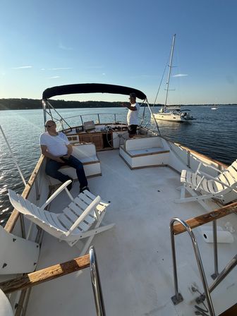 Two men on a motorboat’s aft deck with white lounge chairs and helm under a black bimini, calm coastal harbor waters and anchored sailboats at golden hour