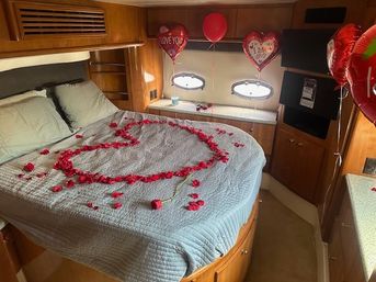 Yacht cabin bedroom decorated for Valentine’s Day with a gray quilted bed, rose petals arranged in a heart, a single red rose, heart-shaped balloons that say I love you, and round porthole windows.