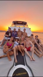 Group of friends lounging on the bow of a white yacht during a golden-orange sunset over calm water — summer sunset yacht cruise on the bay.