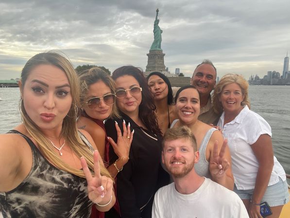 Group selfie on a boat tour in New York Harbor with the Statue of Liberty and Manhattan skyline in the background under a cloudy sky.
