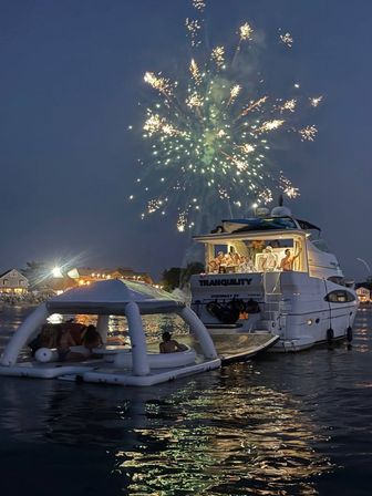 Fireworks burst over a luxury yacht and nearby inflatable floating lounge at dusk, people enjoying a summer boat party with colorful reflections on the water and lit shoreline in the background.
