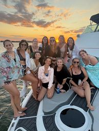 Group of women enjoying a sunset yacht cruise, posing on the boat's bow with drinks, sunglasses and summer blankets against a colorful sky over the water