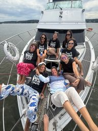 Eight women relaxing on the bow of a white motor yacht, wearing matching event shirts and sunglasses, holding drinks during a summer boat outing on a calm bay under a cloudy sky.
