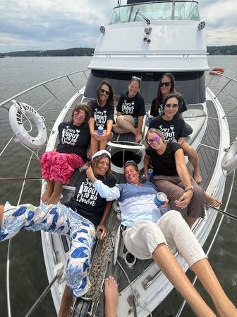 Eight women relaxing on the bow of a white motor yacht, wearing matching event shirts and sunglasses, holding drinks during a summer boat outing on a calm bay under a cloudy sky.