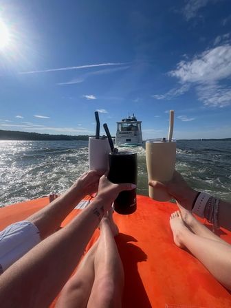 Three people toasting tumblers on an orange float behind a yacht on a sunny summer lake with blue sky and visible boat wake