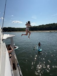 Person mid-air jumping off a boat into a sparkling lake while a paddleboarder watches nearby, sunny summer day with tree-lined shoreline.