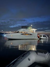 Luxury motor yacht anchored at twilight in a calm harbor, glowing interior lights reflecting on the water and an inflatable dinghy in the foreground under a deep blue sky.