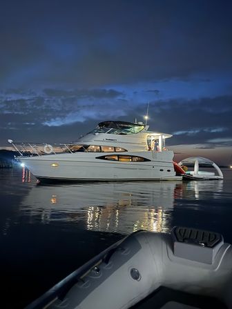 Luxury motor yacht anchored at twilight in a calm harbor, glowing interior lights reflecting on the water and an inflatable dinghy in the foreground under a deep blue sky.