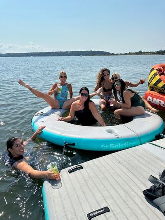 Six friends relaxing on a round inflatable float and dock at a sunny lake, wearing swimsuits, holding drinks and smiling beside a colorful towable tube on a summer outing.