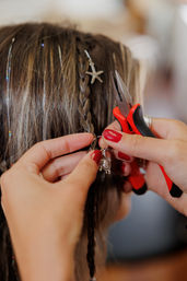 Close-up of hands attaching a silver turtle charm to a small braided hair section with pliers, red-painted nails, starfish charm and hair tinsel in a beachy boho hairstyle.