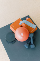At-home workout gear on a dark yoga mat: orange mini exercise ball, two foam yoga blocks, light-blue resistance band, sliding discs and a pair of small dumbbells.
