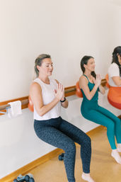 Women in a bright fitness studio doing barre-style wall squats with small stability balls against a barre, palms pressed together in a focused group workout.