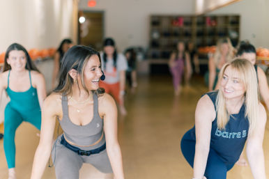 Smiling female instructor wearing a headset leading a lively barre class in a bright fitness studio as participants follow along in lunges