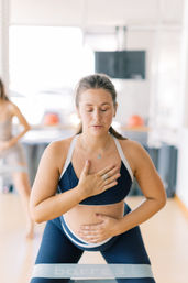 Pregnant woman in navy activewear practicing prenatal breathing with hand on chest and belly, using a resistance band in a bright yoga/fitness studio