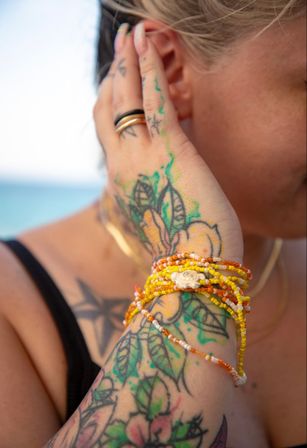 Close-up of a tattooed forearm and hand wearing stacked yellow-orange beaded bracelets and rings, hair tucked behind the ear with a blurred beach and ocean background.