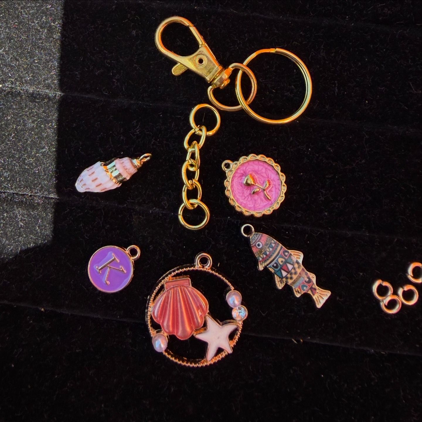 Close-up flat lay of a gold keyring and lobster clasp with colorful beach-themed charms — pink shell and starfish pendant, patterned fish charm and purple initial disc on black velvet background.