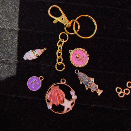 Close-up flat lay of a gold keyring and lobster clasp with colorful beach-themed charms — pink shell and starfish pendant, patterned fish charm and purple initial disc on black velvet background.