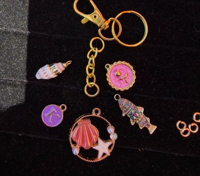 Close-up flat lay of a gold keyring and lobster clasp with colorful beach-themed charms — pink shell and starfish pendant, patterned fish charm and purple initial disc on black velvet background.
