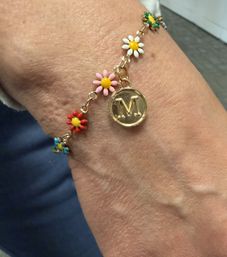 Close-up of a wrist wearing a gold charm bracelet with colorful enamel daisy flowers (red, pink, white, blue) and a round gold monogram 'M' medallion