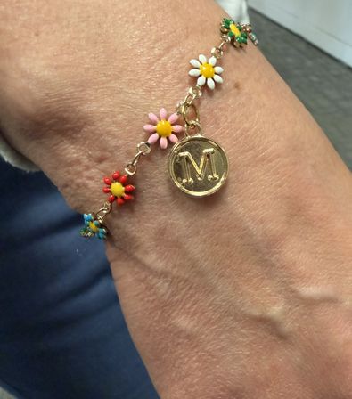 Close-up of a wrist wearing a gold charm bracelet with colorful enamel daisy flowers (red, pink, white, blue) and a round gold monogram 'M' medallion
