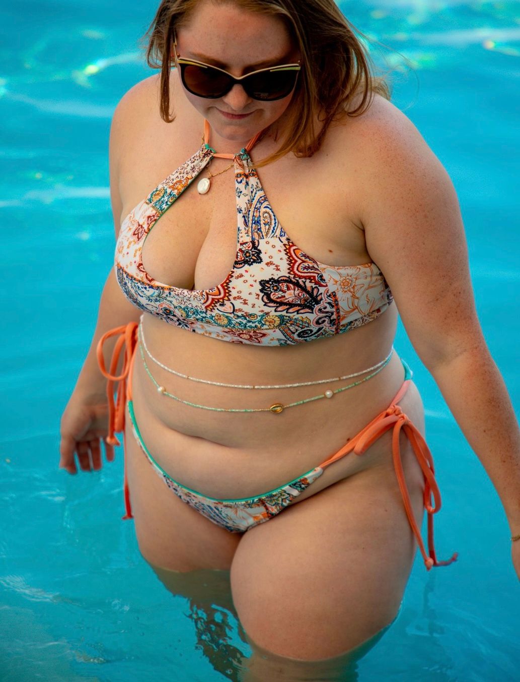 Curvy person in a colorful paisley bikini and sunglasses wading in a bright blue pool, tie-side bottoms and layered waist chains for a playful summer look.