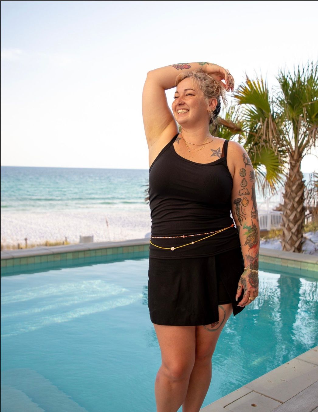 Smiling tattooed woman in a black tank top and skirt posing by a beachfront pool with palm trees and turquoise ocean — coastal beach vacation vibe.