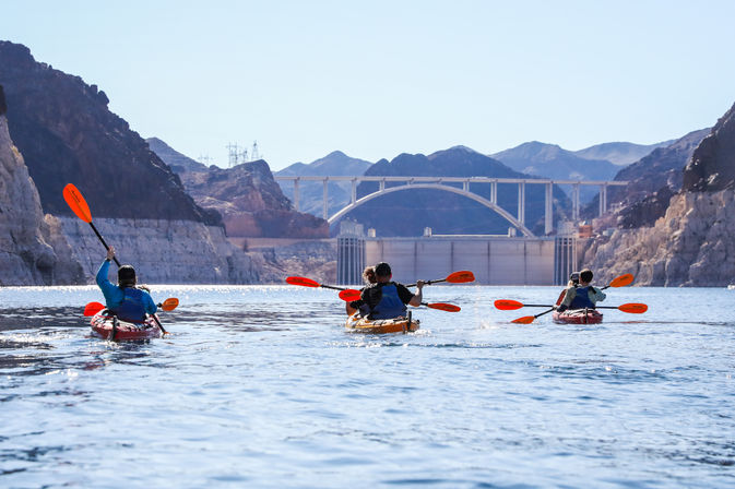 Three kayakers with bright orange paddles gliding on blue Lake Mead toward Hoover Dam and the arched memorial bridge framed by rugged canyon walls
