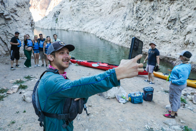 Man taking a selfie with friends on a canyon riverbank during a kayaking trip, colorful kayaks and gear on the shore with steep rocky cliffs rising behind them.