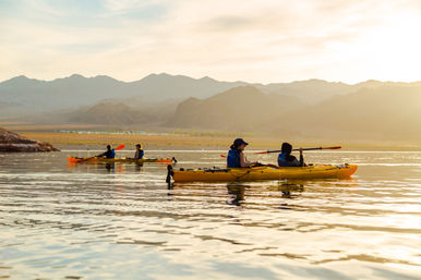 Two tandem yellow kayaks with paddlers in life vests gliding across a calm mountain lake at golden sunset, soft light reflecting on the water