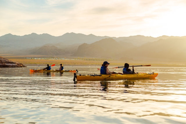 Two tandem yellow kayaks with paddlers in life vests gliding across a calm mountain lake at golden sunset, soft light reflecting on the water
