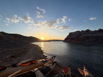 Kayaks pulled ashore on a rocky lake beach at sunset, golden sun reflecting on calm water between rugged hills and cliffs under a blue sky with scattered clouds.