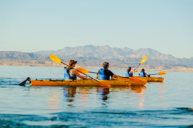 Group of four kayakers in bright yellow kayaks paddling on a calm blue lake with sunlit desert mountains and clear sky reflected in the water