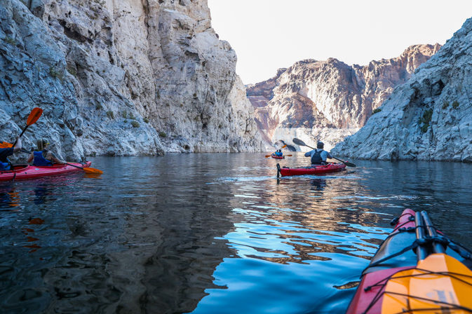 Kayakers paddling red kayaks through a narrow desert canyon on calm reflective blue water, surrounded by towering sunlit limestone cliffs — scenic outdoor kayaking adventure.