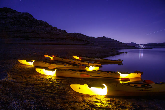 Glowing yellow kayaks lined along a rocky lake shore at night, calm water reflecting distant lights under a starry purple sky.