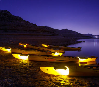 Glowing yellow kayaks lined along a rocky lake shore at night, calm water reflecting distant lights under a starry purple sky.