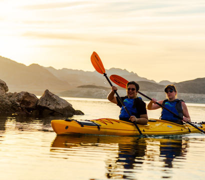 Two people paddling a yellow tandem kayak across a calm mountain lake at sunset, orange paddles glinting in the golden glow with rocky shoreline and silhouetted hills behind them.