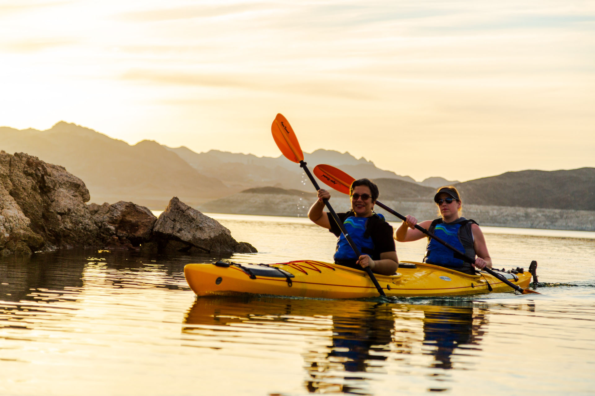 Two people paddling a yellow tandem kayak across a calm mountain lake at sunset, orange paddles glinting in the golden glow with rocky shoreline and silhouetted hills behind them.
