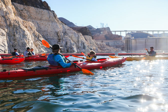 Group of kayakers in red kayaks paddling on a sunlit reservoir below rugged canyon cliffs with a concrete dam and bridge in the background, sparkling water reflecting the sun.