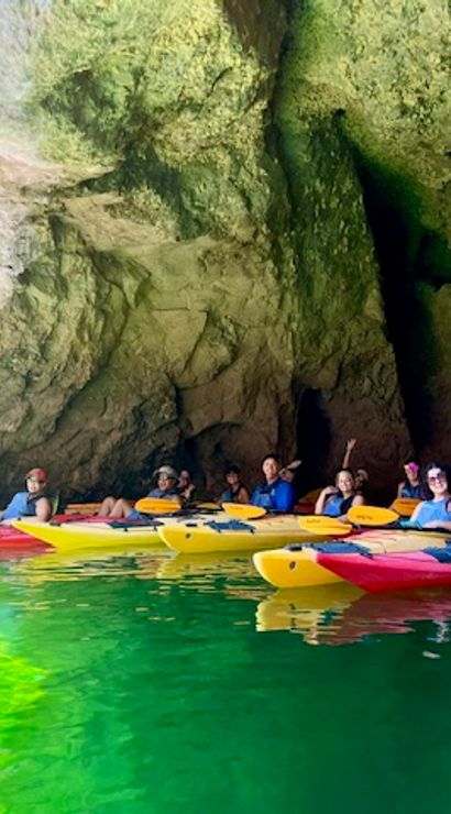 Group of kayakers in colorful kayaks floating inside a sunlit coastal sea cave over emerald-green water