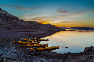 Yellow kayaks lined up on a rocky lakeshore at sunset, calm water reflecting a colorful sky and distant hills