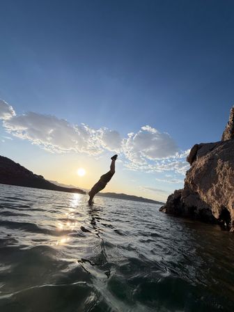 Silhouette of a person diving headfirst into a lake from a rocky cliff at sunset, vertical body reflected in rippling water beneath a blue sky with scattered clouds.