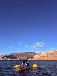 Two people kayaking with yellow paddles on a calm desert lake, framed by red‑rock cliffs and a vast blue sky — scenic canyon paddling adventure.