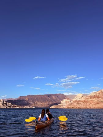 Two people kayaking with yellow paddles on a calm desert lake, framed by red‑rock cliffs and a vast blue sky — scenic canyon paddling adventure.