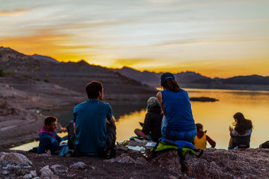 Group of people sitting on a rocky lakeshore watching a golden sunset over mountain ridges, calm water reflecting the colorful sky